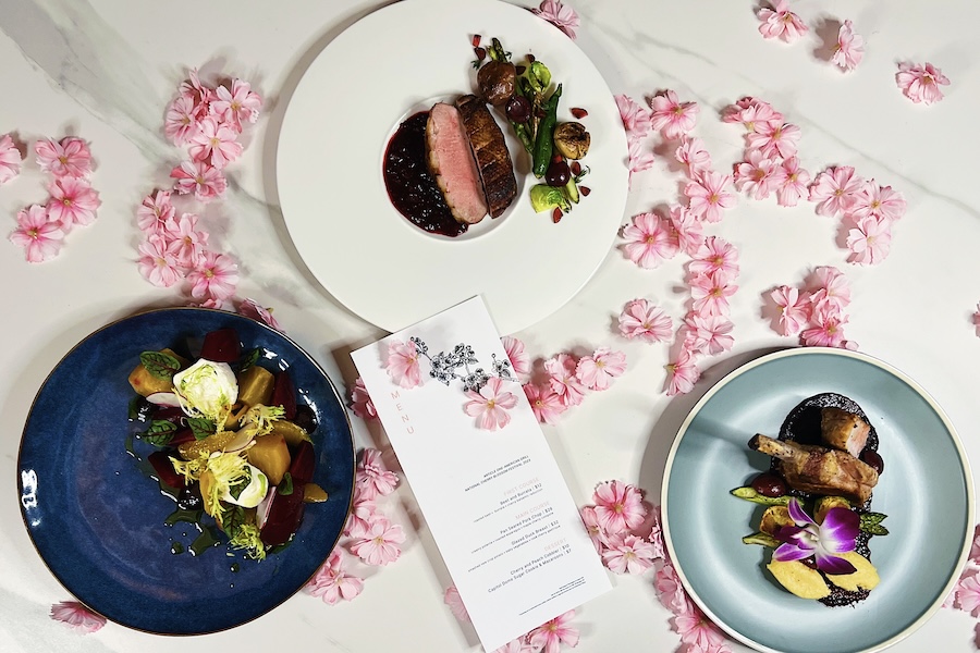 view from above of a table with three plates of fine dining food, a menu, and cherry blossoms scattered on the tablecloth