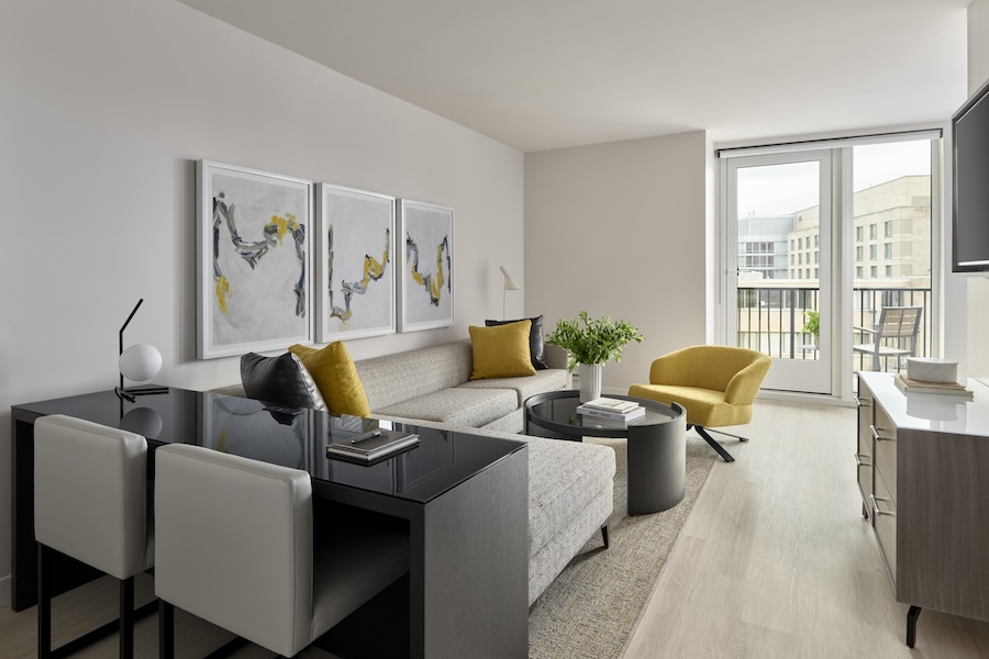 Modern living room with a light gray sectional sofa, yellow accent chair, and black coffee table. Abstract art hangs on the wall, and a large window provides natural light and a view of nearby buildings.