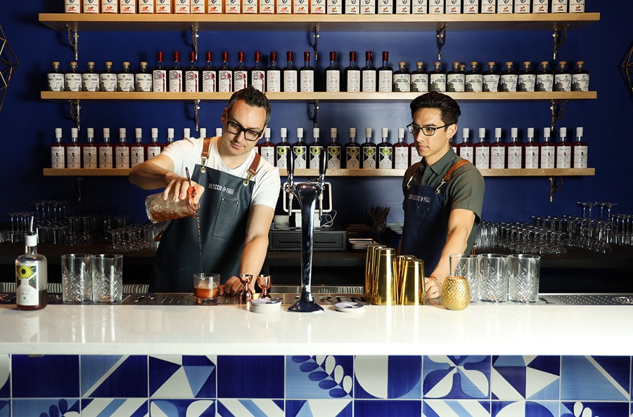 um barman de óculos e avental serve um coquetel enquanto outro observa, com prateleiras de licores nos fundos