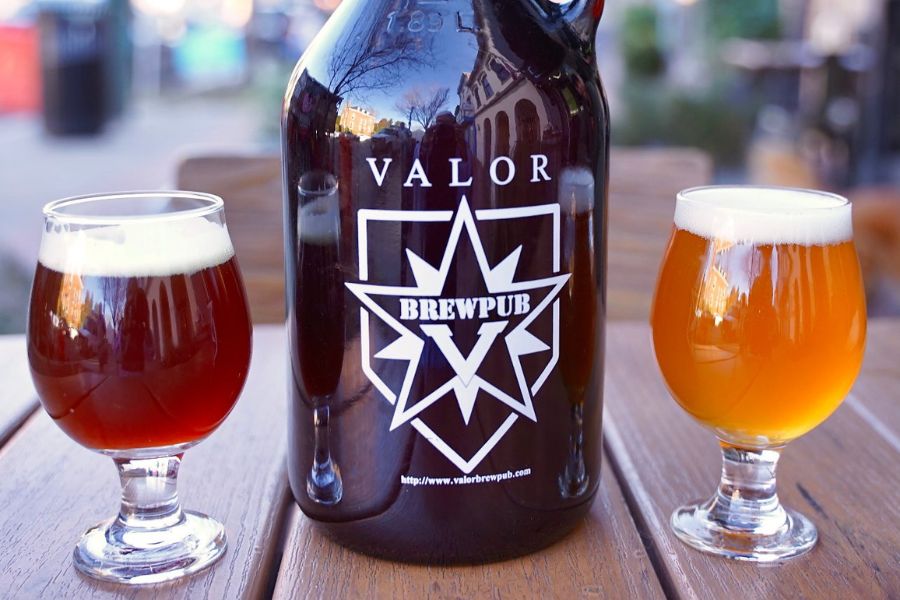 Un growler y dos vasos de cerveza sobre una mesa de madera al aire libre, con el logo de Valor Brewpub. Las cervezas son de color ámbar y dorado pálido.