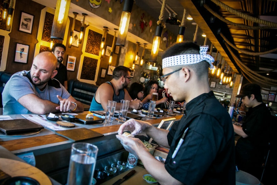  A busy sushi bar with a chef preparing food in the foreground and customers seated along the counter, enjoying their meals.