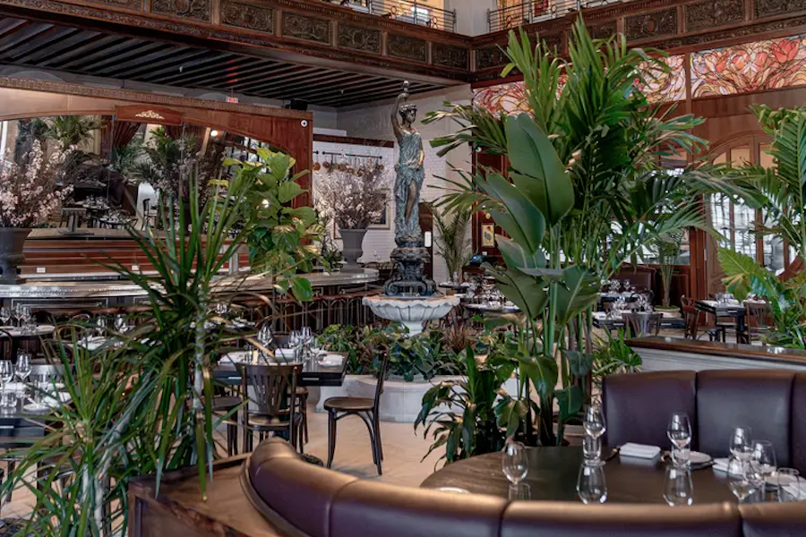 Elegant restaurant interior with a central fountain statue, lush green plants, dark wood furnishings, and tables set with glassware. Stained glass and intricate woodwork adorn the upper walls.