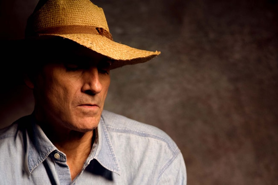 James Taylor wearing a straw hat and a denim shirt, looking down thoughtfully against a dark, muted background.