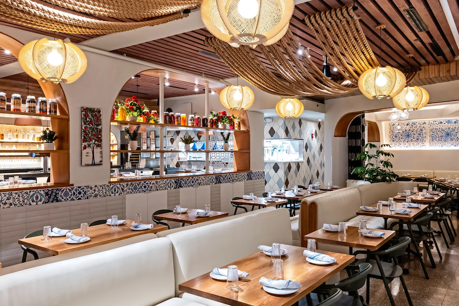 Stylish restaurant interior with wooden tables, white cushioned seating, and decorative lighting. Shelves with colorful jars and plants add to the vibrant, cozy atmosphere. Rope decor hangs from the ceiling.