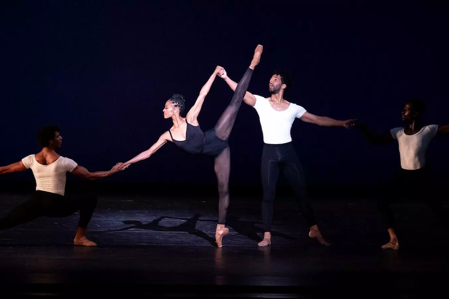 A ballet dancer in a black leotard performs an arabesque on pointe, supported by three male dancers in white shirts and black pants on a dimly lit stage.