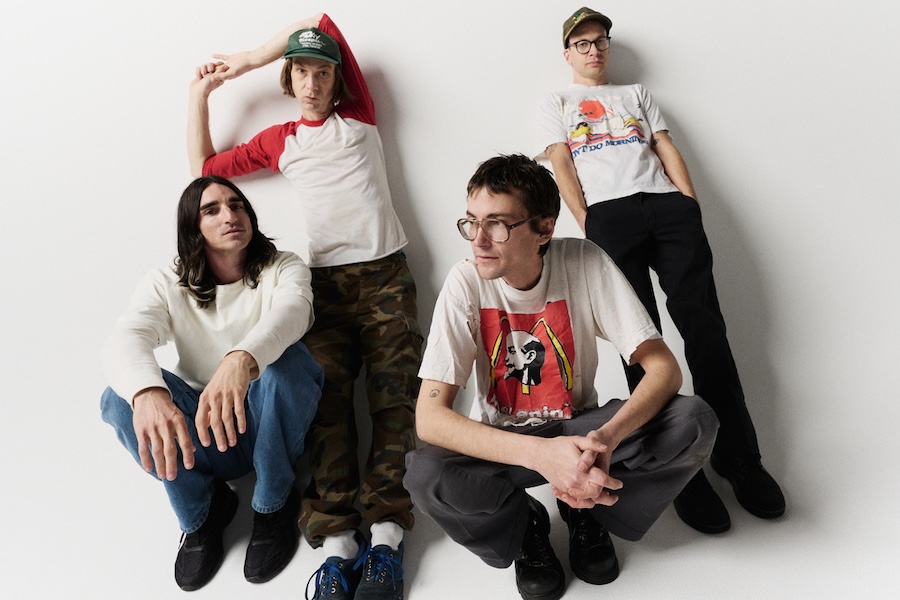 A group photo of four men from a band, sitting and standing against a plain white background. They are casually dressed in t-shirts, jeans, and caps.