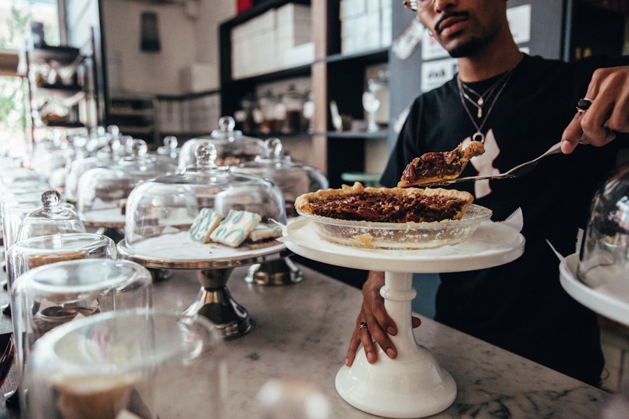 A person serves a slice of pie from a display at a bakery, with a variety of baked goods under glass domes lined up on a marble counter in a cozy, well-lit setting.