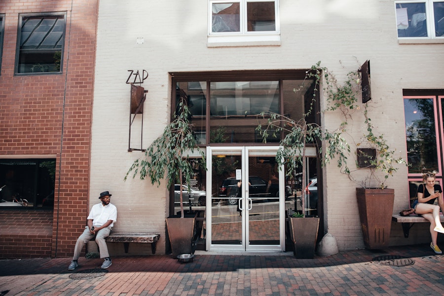 The exterior of a bakery with a sign that reads 'ZAP' above a glass door entrance. Two large potted plants with trailing vines frame the entrance. A man wearing a hat sits on a bench to the left, while a woman sits on a bench to the right, engrossed in her phone.