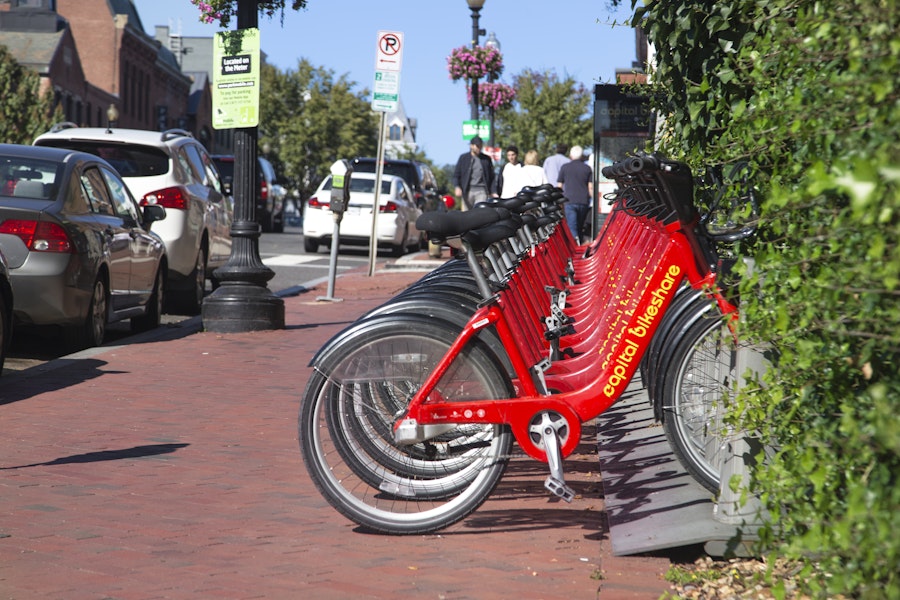 A row of bright red Capital Bikeshare bicycles lined up on a sidewalk in a vibrant city area.