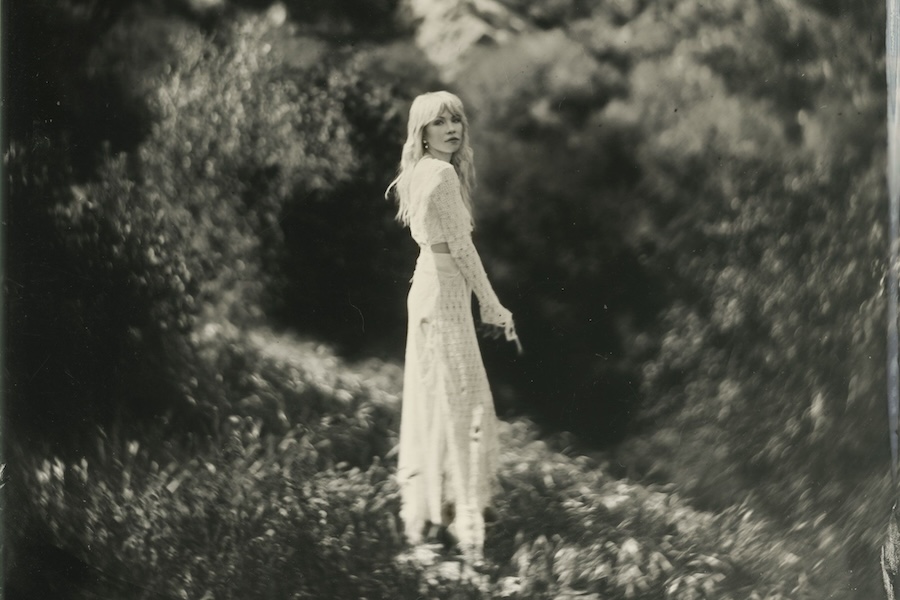 A black and white photo of Carly Rae Jepsen standing in a natural setting, wearing a long, light-colored dress and looking back at the camera.