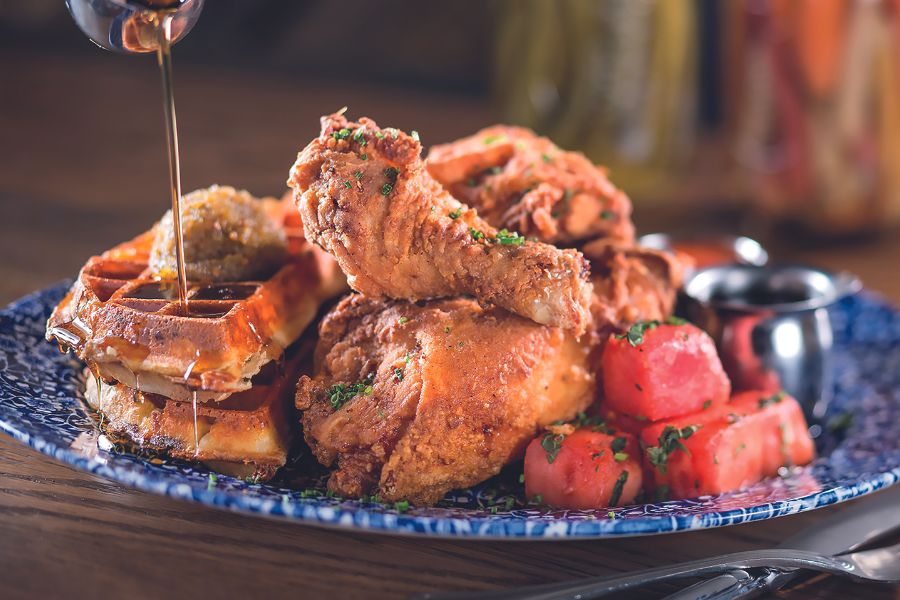  Blue-patterned plate of classic chicken and waffles with crispy fried chicken, golden waffles, syrup being drizzled on top, and watermelon pieces garnished with herbs.