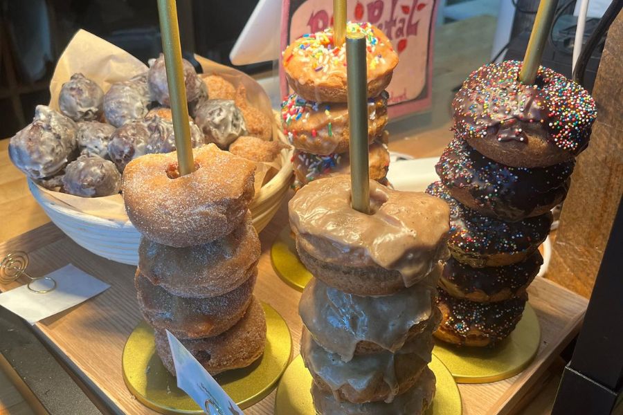 Stacks of various gluten-free donuts on display, with a basket of donut holes in the background. The donuts are arranged on stands, featuring different glazes and sprinkles.