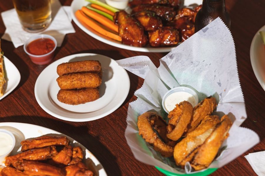 A variety of fried appetizers, including onion rings, mozzarella sticks, and chicken wings. The food is arranged on plates and in baskets, with dipping sauces and vegetable sticks on the side.