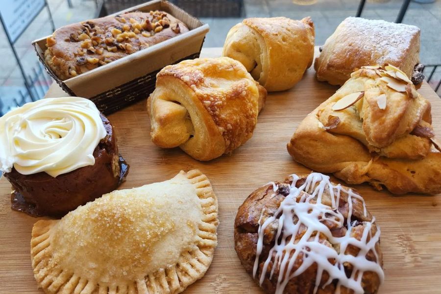 An assortment of baked goods, including pastries, a cinnamon roll with icing, a turnover, and a loaf of bread with nuts. The items are arranged on a wooden board, highlighting their variety and appeal.