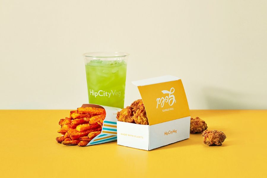 This image showcases a meal from HipCityVeg, featuring a box of plant-based fried chicken, a serving of sweet potato fries, and a green drink in a branded cup. The items are displayed on a yellow surface, emphasizing the vibrant and appealing presentation.