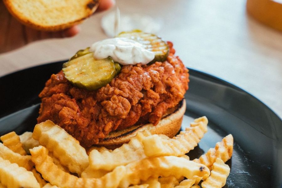 A vegan fried chicken sandwich topped with pickles and a dollop of sauce, served with crinkle-cut fries on a black plate. The sandwich bun is being held above the sandwich, showcasing the ingredients.