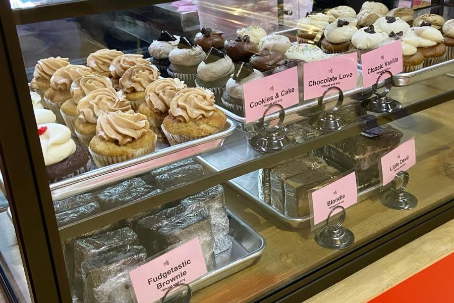 a display case filled with a variety of vegan cupcakes and baked goods. The cupcakes, labeled with flavors like "Cookies & Cake," "Chocolate Love," and "Classic Vanilla," are neatly arranged on trays. Below the cupcakes, there are brownies and other treats, including "Fudgestastic Brownie" and "Blondie."