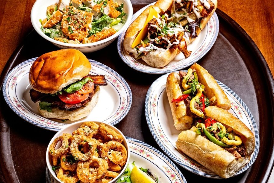 This image showcases a variety of vegan dishes, including a burger, two sandwiches, a salad, and a plate of onion rings. Each dish is served on a decorative plate, adding to the appetizing presentation. 