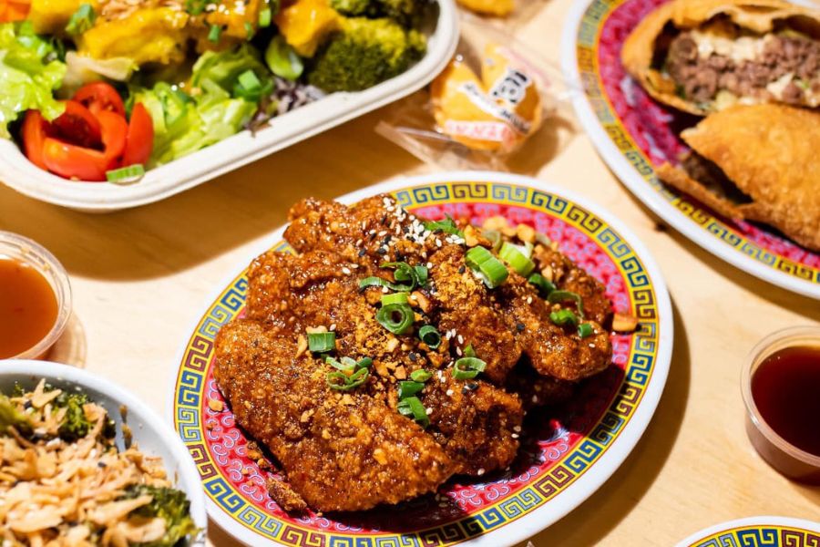 This image features a plate of crispy, sauced chicken garnished with green onions and sesame seeds. Surrounding the main dish are various other items, including a salad, dumplings, and dipping sauces, all arranged on a table. 