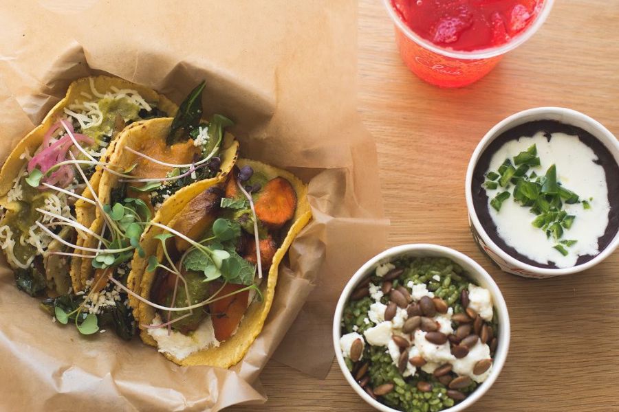 This image showcases a serving of three tacos filled with various vegetables and garnished with greens, placed on brown paper. Alongside, there are bowls of black beans, a green rice dish with seeds and cheese, and a red drink, all arranged on a wooden surface.
