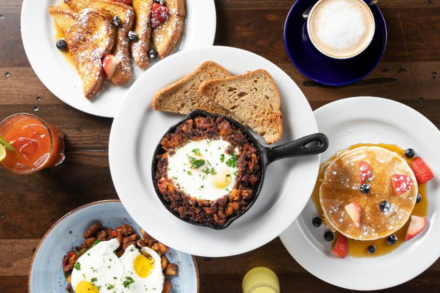 This image displays a variety of breakfast dishes, including French toast with berries, a skillet dish with eggs and toast, a stack of pancakes with fruit, and a plate of hash topped with eggs. There is also a cappuccino and a Bloody Mary, all arranged on a wooden table.
