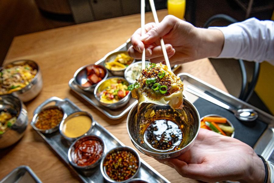 This image features a person using chopsticks to dip a dumpling into a flavorful sauce. In the background, there are various bowls and trays filled with different dishes and condiments, showcasing a diverse and appetizing meal spread.