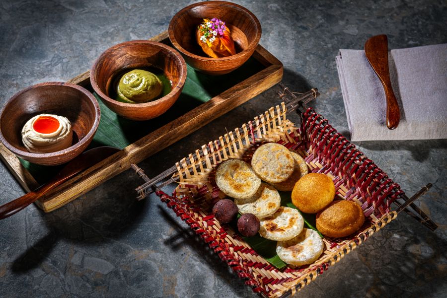 This image features an assortment of arepas and spreads arranged in a woven basket and wooden bowls. The setup is complemented by wooden utensils and a napkin on a textured surface.