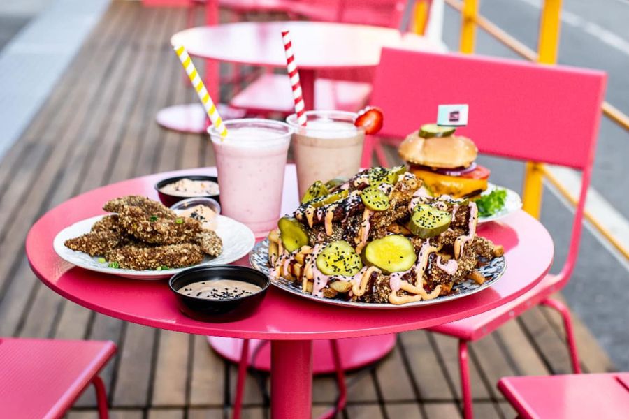 A vibrant pink table set with a delicious spread of plant-based food, including crispy chicken-style tenders, loaded fries topped with pickles and drizzled sauce, creamy milkshakes, and a juicy burger. 