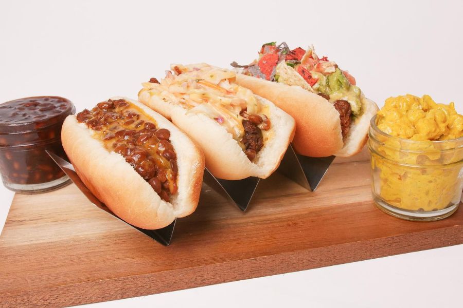 Three plant-based hot dogs are topped with various delicious ingredients, accompanied by small jars of baked beans and macaroni and cheese on a wooden board.
