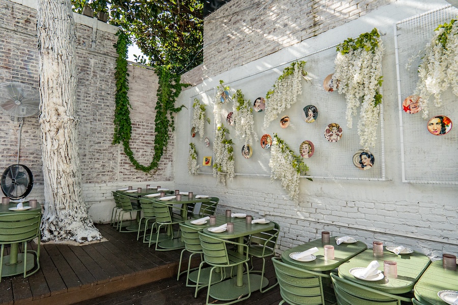 Outdoor dining area with green tables and chairs, decorated with hanging vines, white flowers, and plates featuring portraits. A large painted tree trunk is on the left.