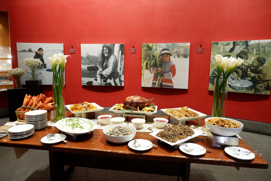Buffet table set with a variety of dishes including a roasted turkey, salads, and sides, set against a red wall decorated with large photographs of people. Calla lilies in tall vases add a touch of elegance to the setting.