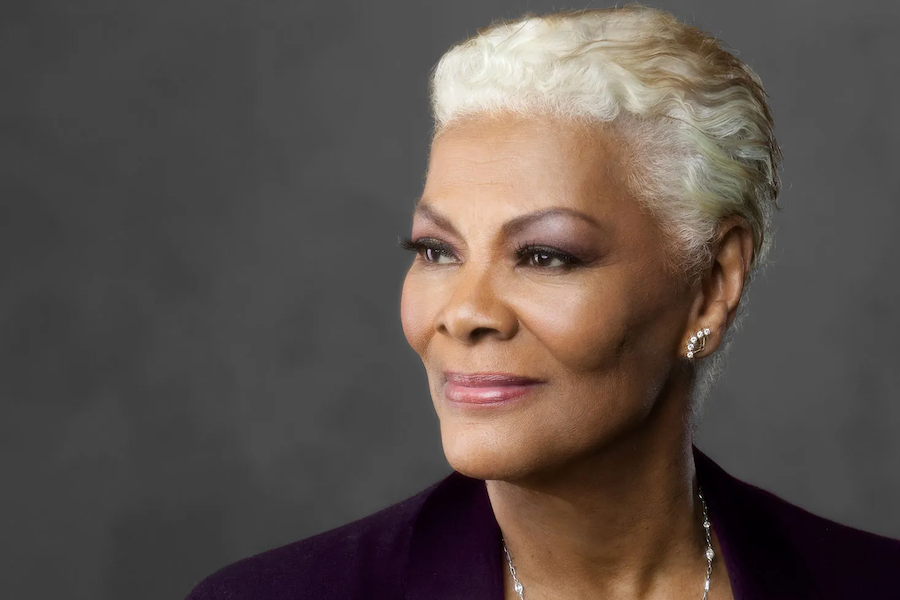 A close-up portrait of Dionne Warwick, with short blonde hair and a calm expression, wearing a dark top against a gray background.