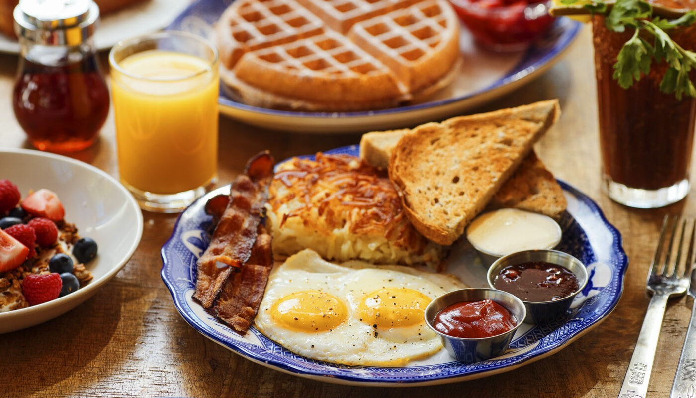 A breakfast plate with eggs, bacon, toast, and hash browns, accompanied by a glass of orange juice, yogurt with berries, and a waffle in the background.