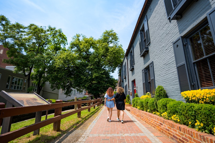 Two women walking along a brick pathway next to a historic canal, flanked by green trees and buildings.