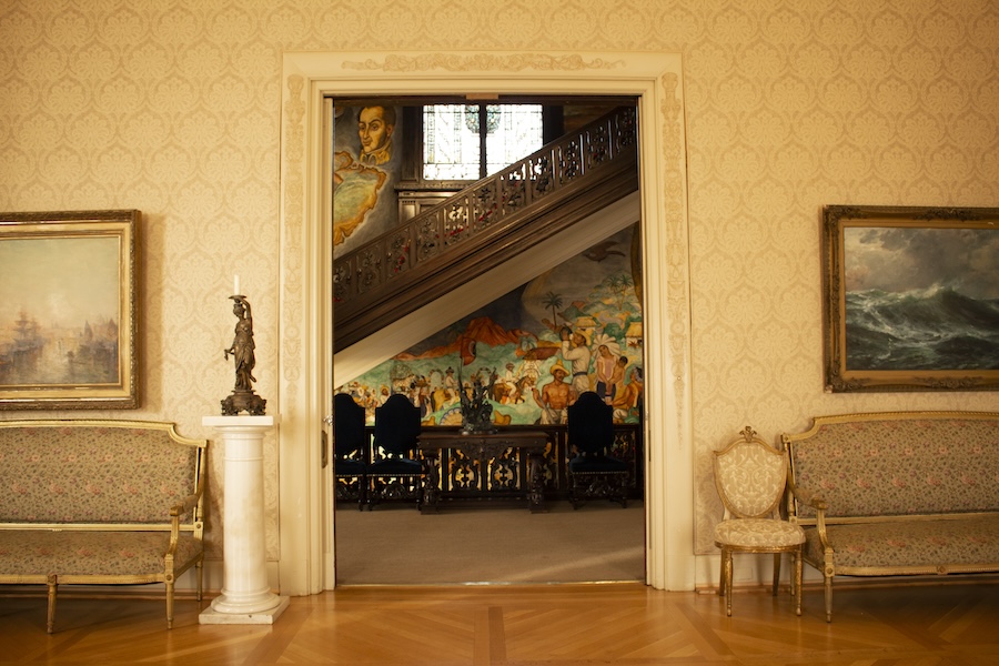 A grand interior space with elegant furniture, framed artwork on the walls, and a view through an open doorway revealing a colorful mural and an ornate staircase.