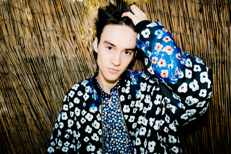 A young man with dark hair wearing a floral patterned jacket and shirt, standing in front of a bamboo background.