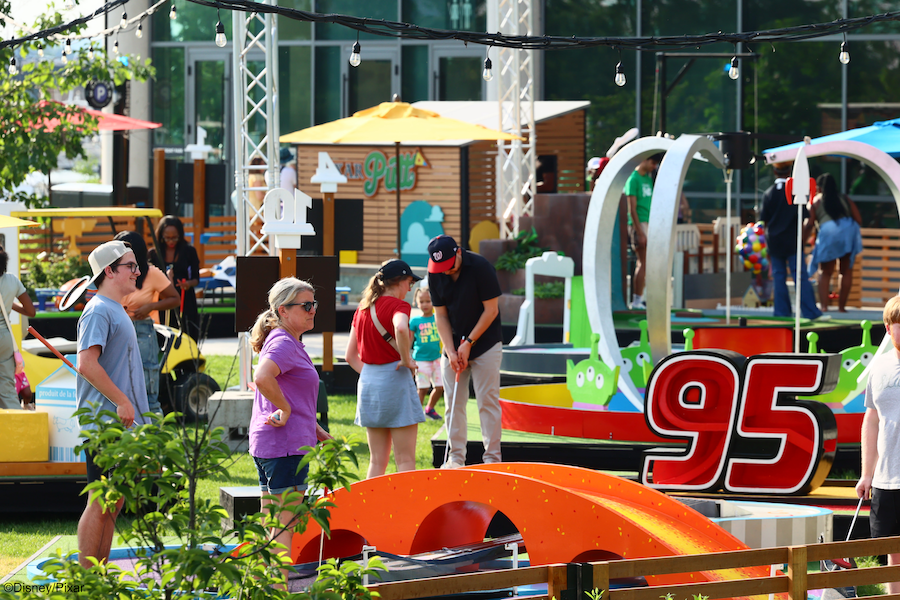People enjoying an outdoor mini golf course with colorful obstacles and decorations themed around Pixar films, including a large number 95 from Cars. The area is lively with participants of various ages.