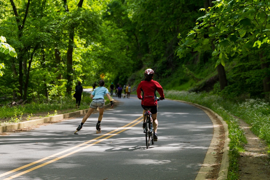 People enjoying a sunny day at Rock Creek Park, with a cyclist and rollerblader on the path surrounded by lush green trees.