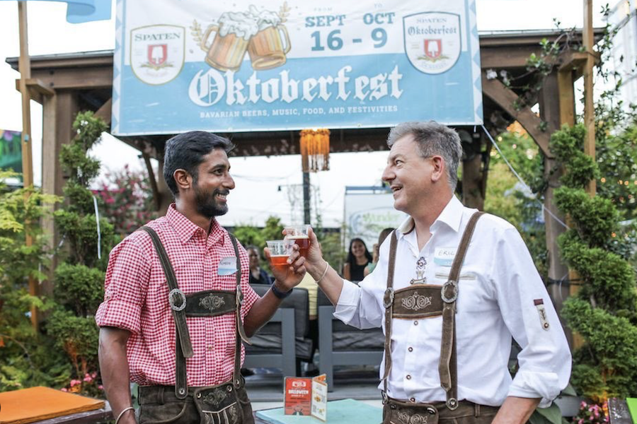 Two men in traditional Oktoberfest attire clinking glasses of beer at an outdoor Oktoberfest event.