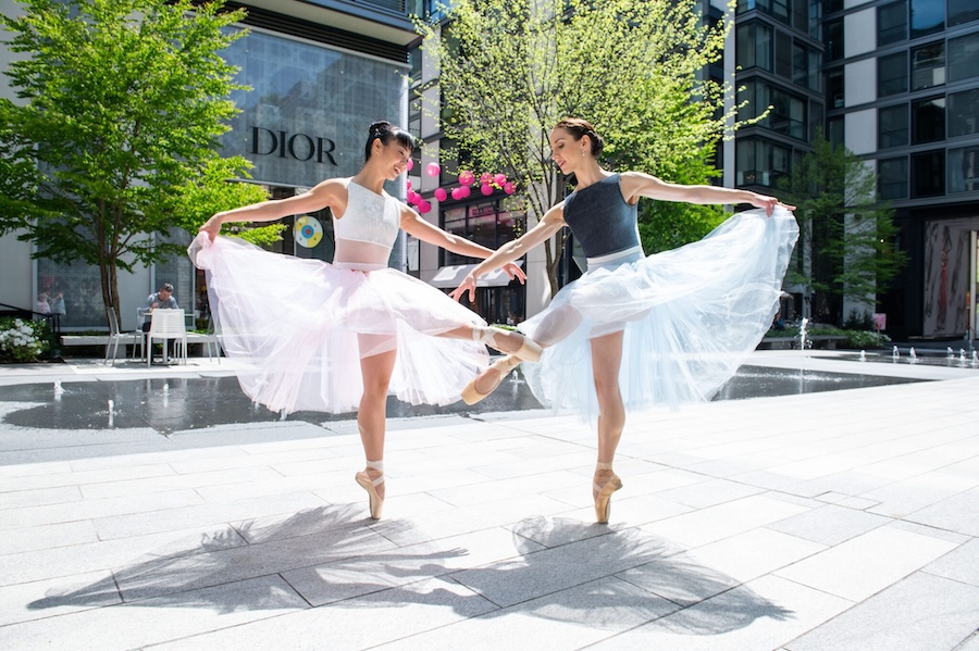 Two ballet dancers in pastel-colored tutus perform an elegant dance move in an outdoor urban setting, with a Dior store in the background and a modern plaza featuring trees and a water fountain.