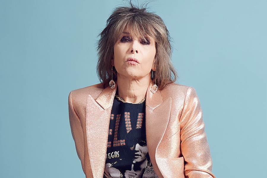A portrait of Chrissie Hynde from The Pretenders, wearing a shiny pink blazer and a black t-shirt with an Elvis print, against a light blue background.