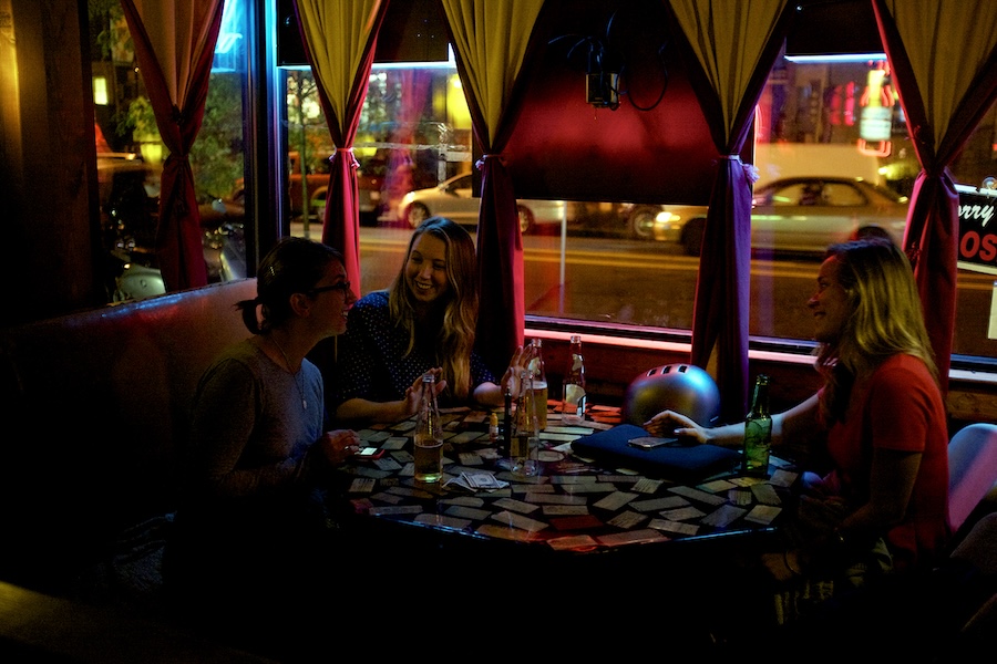 Three friends enjoy a night out, seated at a cozy, dimly lit table with drinks, against a backdrop of vibrant city lights seen through large windows.