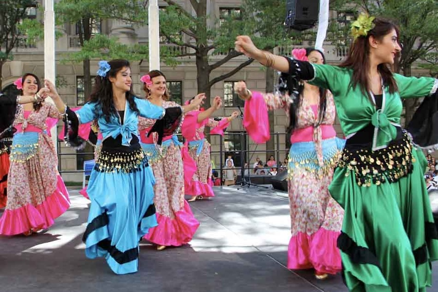 A group of women performing a Turkish dance on stage, wearing colorful traditional dresses with bright blue, green, and pink accents.