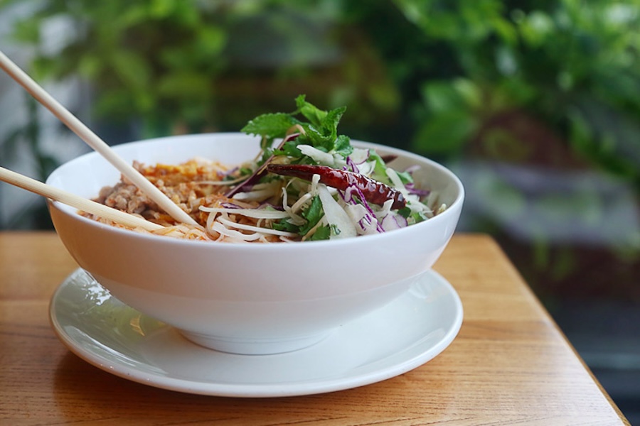 Bowl of Laotian food garnished with fresh herbs and vegetables, with chopsticks placed on the side, set on a wooden table with a green, leafy background.