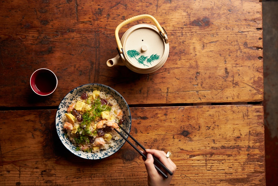 A bowl of rice with vegetables and meat, chopsticks, a teapot, and a small cup on a wooden table.