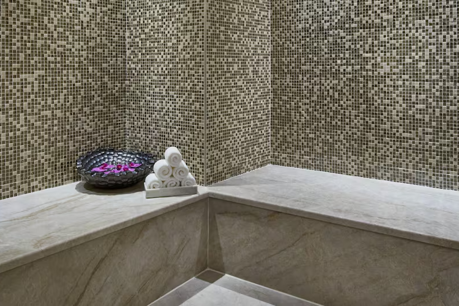 Marble steam room with mosaic tile walls, a black bowl with petals, and rolled towels.