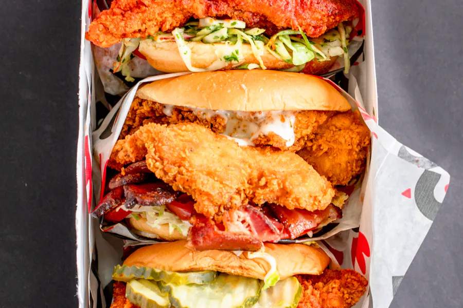 A box of three Roaming Rooster fried chicken sandwiches, each with different toppings, including pickles, coleslaw, and bacon.