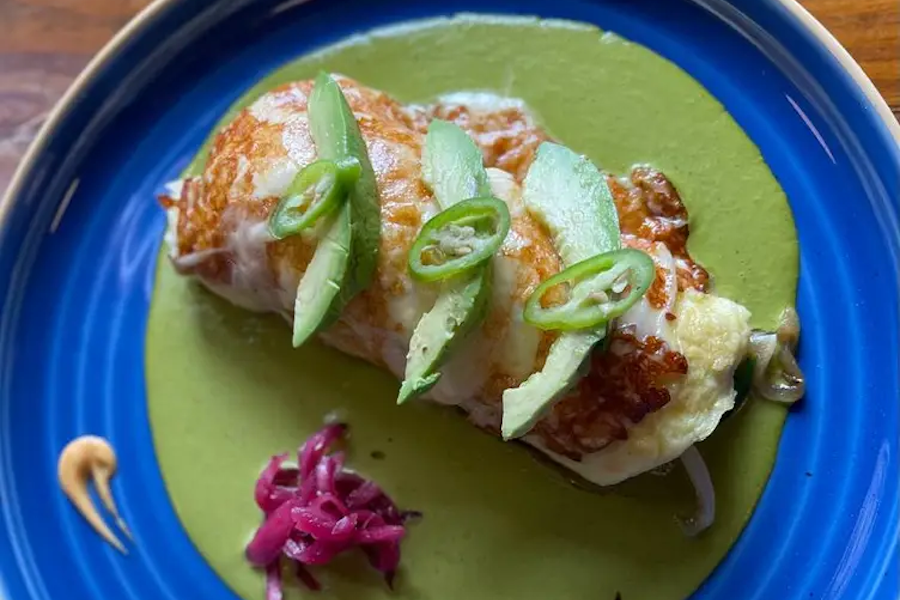 A Mexican dish on a blue plate featuring a stuffed and baked tortilla with melted cheese, topped with sliced green peppers, and served with a green sauce and pickled red onions.