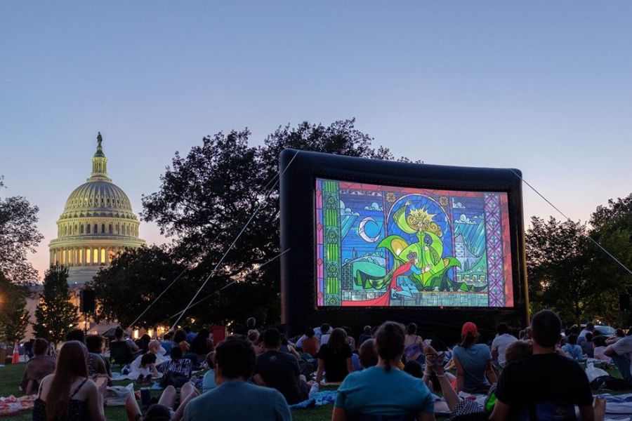 Outdoor movie night at the Library of Congress, featuring an inflatable screen showing a colorful animated film, with a crowd seated on the lawn enjoying the evening under a clear, twilight sky and the U.S. Capitol in the background.