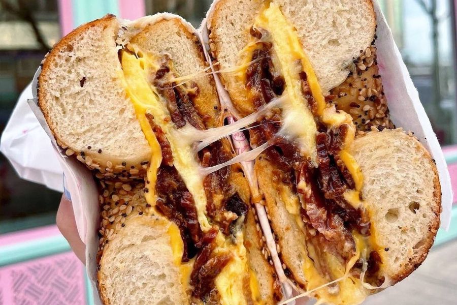 Close-up of a Sun City breakfast sandwich from Call Your Mother, featuring an everything bagel filled with egg, melted cheese, hot honey and bacon.